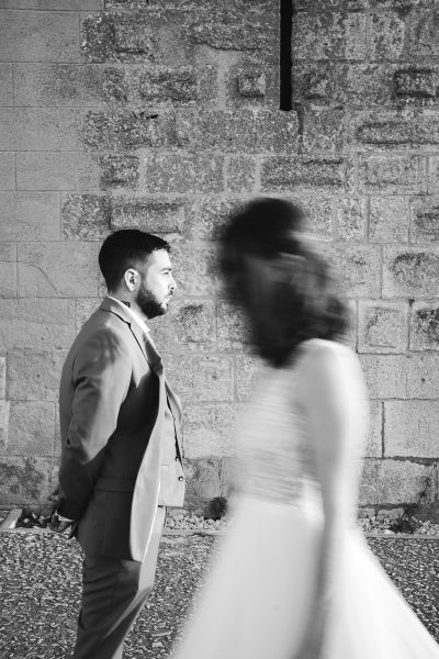 A groom standing in profile against a stone wall while the bride walks past, blurred by movement